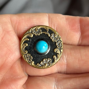 May include: A circular button featuring a central bright turquoise cabochon set against a dark, textured background. The outer edge is framed by an ornate, raised brass-toned metal border decorated with embossed floral and scrolling leaf motifs.