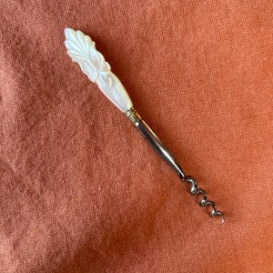 May include: A corkscrew featuring a white iridescent mother-of-pearl handle carved with a leaf-like pattern. The handle connects to a slender metallic shaft that transitions into a dark, spiraled metal worm for extracting corks from bottles.