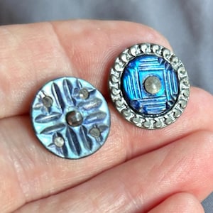 Può includere: Due bottoni circolari con superfici blu e viola iridescenti. Il bottone a sinistra presenta un motivo in rilievo a forma di stella con piccoli accenti metallici. Il bottone a destra ha un centro blu intenso con un bordo metallico e un perno centrale.