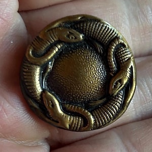 May include: A round, antique brass button featuring a detailed design of several snakes encircling a textured center. The snakes have visible scales and are arranged in a circular pattern. The button has a vintage aesthetic.