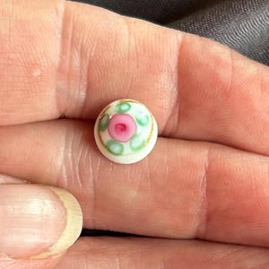 May include: A small, round, white ceramic button featuring a hand-painted floral design. The center displays a pink rosebud surrounded by green leaf accents, all encircled by a thin, delicate gold-colored rim painted along the outer edge.