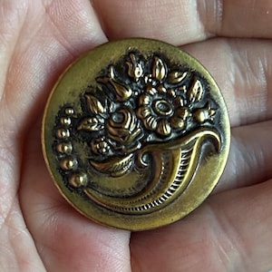 May include: A round, antique brass button with a floral design. The button features a detailed relief of flowers and leaves spilling from a cornucopia. The button is approximately 3.8 cm in diameter.