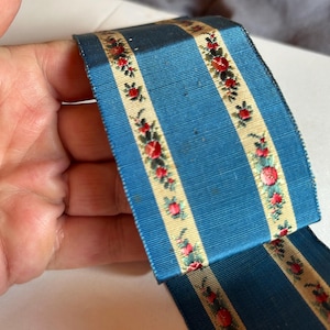 May include: A wide fabric ribbon featuring alternating vertical stripes of solid blue and cream. The cream stripes are decorated with a repeating floral pattern of small red roses and green leaves. The ribbon has a textured, woven appearance.