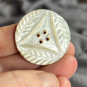 May include: A round, iridescent mother-of-pearl button featuring a central triangular motif with four sewing holes. The surface is decorated with intricate, carved leaf patterns arranged in a geometric layout, showcasing a natural pearlescent sheen.
