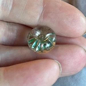 May include: A small, round, clear glass bead featuring a central hole and internal green, ribbed, or fluted detailing that radiates from the center toward the outer edge, creating a textured appearance within the transparent material.
