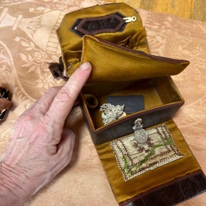 May include: A dark brown leather sewing box with a mustard yellow satin interior. The box features a hinged lid, a metal latch, and an internal needle book with embroidered details. Inside, there is a small piece of white lace and a black rectangle.