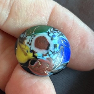 Op de afbeelding: Een ronde glazen knoop met een veelkleurig bloemmotief met wervelingen van geel, blauw, groen en bruin. Het oppervlak is versierd met dunne, metallic goudkleurige lijnen die de organische vormen in het glasontwerp accentueren.