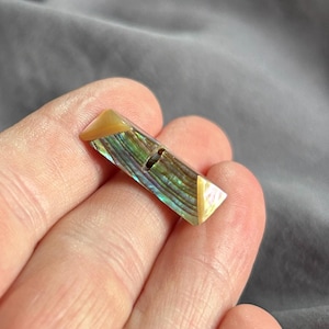 Pode incluir: Um botão retangular tipo alamares com uma incrustação central de concha de abalone iridescente em tons de verde e azul, ladeado por extremidades triangulares lisas de cor clara. O botão possui uma única ranhura horizontal central para passar.