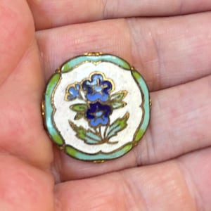 May include: A round metal button featuring white enamel in the center with a painted floral design of blue flowers and green leaves. The outer edge of the button is decorated with a scalloped border of light green enamel and gold-toned metal.