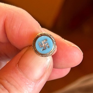 May include: A small circular lapel pin featuring a light blue enamel center with a silver-toned swastika symbol. The pin has a textured, beaded metallic border around the edge, providing a distinct contrast to the smooth, colored inlay.