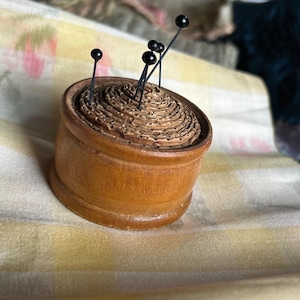 Könnte beinhalten: Ein rundes Nadelkissen aus Holz mit einer strukturierten, spiralförmigen Oberfläche. Mehrere gerade Stecknadeln mit kleinen, runden schwarzen Köpfen stecken in der Mitte des Kissens und ragen in verschiedenen Winkeln aus der Holzbasis heraus.