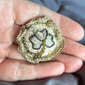 May include: A circular brooch featuring a central embroidered three-leaf clover in white and gold thread on a cream fabric background. The edge is finished with a textured gold braided trim and small silver-toned metallic beads.
