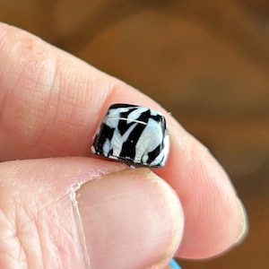 May include: A small, square bead with a black and white zebra stripe pattern. The bead is held between two fingers, showcasing its size and design. The bead's surface appears glossy, reflecting light.
