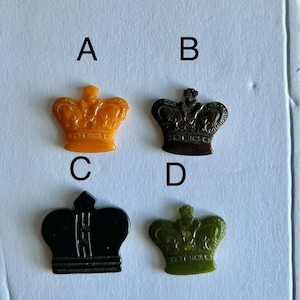 Può includere: Quattro bottoni a forma di corona etichettati A, B, C e D. Il bottone A è arancione, B è marrone scuro, C è nero con fessure verticali e D è verde oliva. Ogni bottone presenta un design a corona dettagliato con fori visibili per la cucitura.