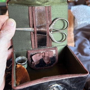 May include: A dark brown sewing box with a green fabric interior. The lid features a pair of silver-toned metal scissors with an ornate handle held by a brown strap. Inside, a small silver thimble and a brown velvet pincushion are visible.