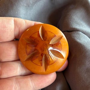 Puede incluir: Un botón redondo de color ámbar con un patrón de estrella de seis puntas tallado que irradia desde el centro. La superficie es lisa y brillante, con ranuras profundas y anguladas que forman el diseño de estrella sobre el material naranja translúcido.