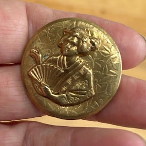 May include: A circular, antique gold button featuring a detailed relief of a person holding a fan. The button has a textured background with floral patterns. The figure is wearing traditional clothing.