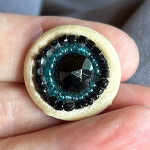 Op de afbeelding: Een ronde knoop met een centrale gefacetteerde zwarte steen, omringd door een ring van kleine turquoise kralen en een buitenrand van piepkleine zwarte kraaltjes. Het geheel is bevestigd op een gladde, licht crèmekleurige stoffen ondergrond.