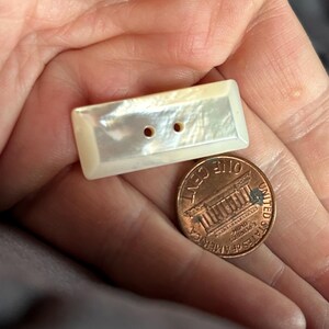 May include: A rectangular, off-white mother-of-pearl button with two center holes, featuring a beveled edge and an iridescent surface, positioned next to a copper-colored United States one-cent coin for a clear visual size reference.