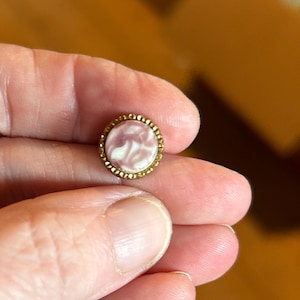 May include: A small circular button featuring a smooth, marbled pink and white center inlay surrounded by a textured, scalloped gold-toned metal rim. The button has a polished finish and a distinct border pattern that frames the central color swirl.