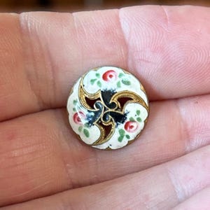 May include: A small round button featuring a white enamel surface decorated with hand-painted red rosebuds and green leaves. The center of the button has a gold-toned metal cutout design in a swirling triskelion pattern against a black background.