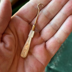 May include: A button hook featuring a slender, metallic copper-toned shaft with a curved hook at the top and a smooth, pearlescent white handle with a marbled texture. The tool is designed for fastening buttons on clothing and accessories.