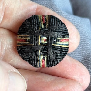 May include: A circular button featuring a woven design of black thread layered over accents of red, yellow, and blue fibers. The surface displays a grid-like pattern created by the interlacing of dark threads, revealing small pops of color beneath.