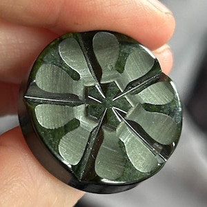 May include: A circular dark green button featuring a metallic, silver-toned inlay pattern that resembles a six-petaled flower or a starburst design. The surface of the button is smooth and glossy, with a distinct contrast between the dark green base and the reflective, brushed silver-colored metal segments embedded into the top face.