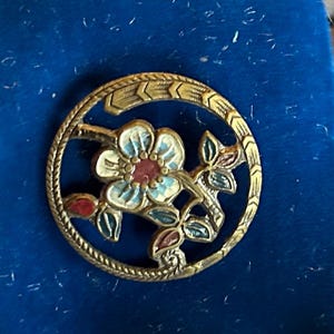 May include: A circular gold-tone metal brooch featuring an openwork design with a central flower. The flower has white petals with blue shading and a red center, surrounded by green and red leaves. The outer rim has a textured rope pattern.