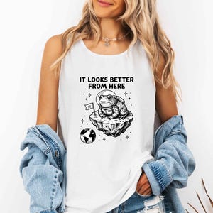 May include: A white sleeveless tank top featuring a black graphic print of a toad wearing an astronaut helmet, sitting on a floating rock with a small flag. The text above the graphic reads IT LOOKS BETTER FROM HERE, with a small Earth icon below.