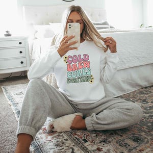 May include: A white crewneck sweatshirt featuring the text COLD BREW CURES EVERYTHING in stacked, colorful, wavy lettering. The design includes small yellow and white daisy accents around the words. The sweatshirt has long sleeves and a ribbed hem.