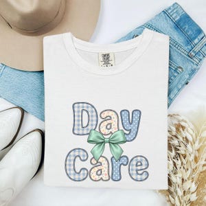 Może przedstawiać: Biały t-shirt z krótkim rękawem z napisem Day Care w zabawnym kroju pisma. Litery wypełnione są niebieską kratką vichy, wzorami kwiatowymi i kropkami. Duża, jasnozielona kokarda jest wyśrodkowana pomiędzy słowami Day i Care.