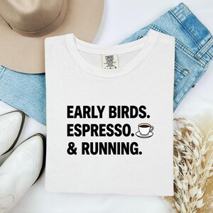 Può includere: Una t-shirt bianca con testo nero che recita EARLY BIRDS. ESPRESSO. & RUNNING. accanto a una piccola grafica di una tazzina da caffè su un piattino. La maglietta presenta un'etichetta del marchio Comfort Colors sul colletto interno.