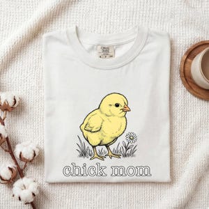 Puede incluir: Camiseta blanca con una ilustración de pollito amarillo y el texto "chick mom". El pollito está de pie en la hierba junto a una margarita. La camiseta es de material suave.