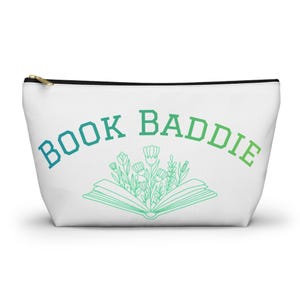 May include: White zippered pouch with the text "BOOK BADDIE" in a gradient blue to green font. Below the text is an open book illustration with flowers and leaves in a similar gradient color.