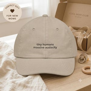 Peut inclure: Une casquette de baseball beige avec le texte brodé 'tiny humans massive audacity' en fil marron foncé. La casquette présente une calotte structurée, une visière incurvée avec des détails surpiqués et un bouton sur le dessus.