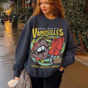 Puede incluir: Una sudadera gris carbón de cuello redondo con un estampado gráfico que incluye el texto VAMPSIGLES, WELL-BITTER READERS SOCIETY, LOVE AT FIRST BITE y SUPPORT YOUR LOCAL VAMPIRES. El diseño muestra unos labios con colmillos y un polo rojo.
