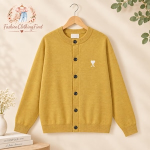 May include: A mustard yellow long-sleeve knit cardigan featuring a round neckline, a front button closure with dark buttons, and a small white heart-shaped logo embroidered on the left chest. The garment is displayed on a wooden hanger.