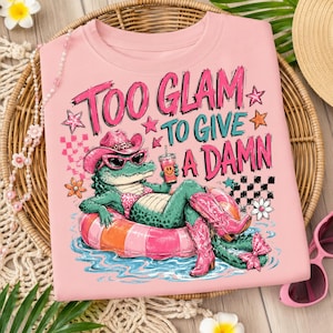 Peut inclure: T-shirt rose avec un motif d'alligator portant un chapeau de cow-boy, des lunettes de soleil et des bottes, se prélassant sur un flotteur de piscine. Le texte dit "Too Glam To Give A Damn". L'alligator tient une boisson.
