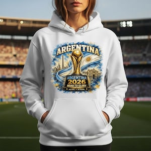 Op de afbeelding: Een witte hoodie met een grafische print van een gouden voetbaltrofee, de Argentijnse vlag en de tekst ARGENTINA 2026 ROAD TO GLORY UN PAIS UN SUE&Ntilde;O. Het ontwerp bevat architecturale silhouetten en een blauw en geel verfspat-effect.