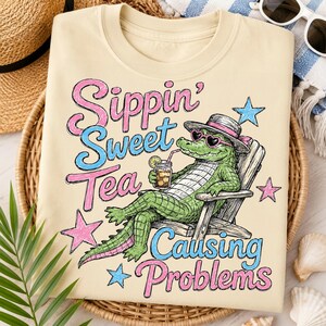 May include: Cream-colored t-shirt with a graphic of an alligator in a beach chair, wearing sunglasses and a hat, sipping iced tea. Text reads "Sippin' Sweet Tea Causing Problems" in pink and blue.