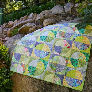 May include: A vibrant quilt with a circular design, showcasing green, blue, and floral fabrics. The quilt is displayed on a large rock outdoors, with a green edge. The design incorporates diverse fabric patterns and textures, creating a patchwork effect.