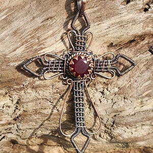 May include: Copper wire-wrapped cross pendant with a central, faceted, red gemstone. The cross features intricate wire weaving and a copper chain. The pendant is displayed on a piece of weathered wood, highlighting the handcrafted details.