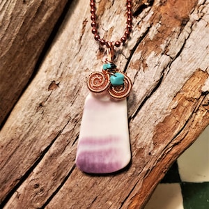 May include: A handcrafted necklace featuring a pendant with a white and purple stone. The pendant is accented with copper wire swirls and turquoise stones. The necklace has a copper chain and is displayed on a piece of driftwood.