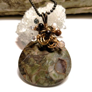 May include: A green, brown, and white marbled stone pendant with a wire-wrapped design featuring black, brown, and white beads. The pendant is attached to a black chain.
