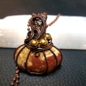 May include: A copper wire wrapped pendant featuring a brown and white agate stone with gold beads. The pendant is attached to a copper chain.