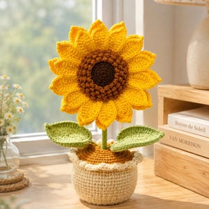 Puede incluir: Un girasol tejido a ganchillo en una maceta. El girasol tiene pétalos amarillos brillantes, un centro marrón y hojas verdes. La maceta beige tiene un borde festoneado, creando una pieza decorativa encantadora.