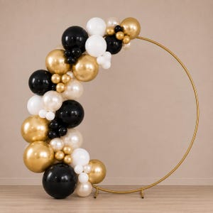 May include: A large circular gold metal frame decorated with a balloon arch. The arrangement features a mix of black, metallic gold, white, and pearlescent cream balloons of varying sizes, clustered along the left side of the circular stand.