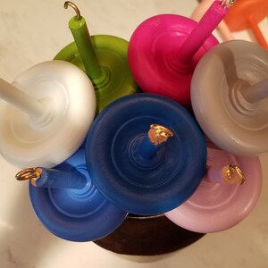 以下が含まれることがあります： 金色のアクセントが付いたカラフルな木製糸巻きコレクション。糸巻きは円形に配置されており、青、緑、ピンク、白、グレーなど、さまざまな色があります。