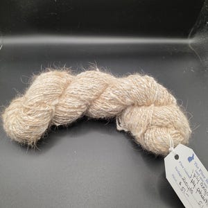 May include: A skein of natural-colored hand-spun yarn. The yarn is a light beige color with a slightly textured appearance. A white tag is attached, with text that includes "The Fiber K", "hand spun yarn", and "ply angora".